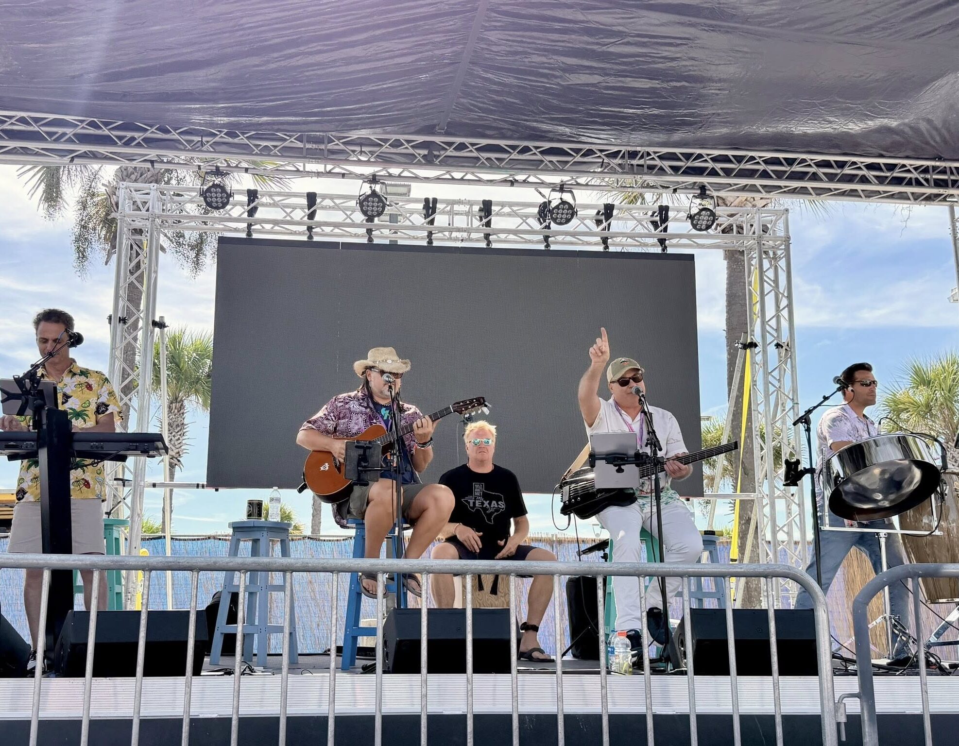 1 Outdoor band performing onstage with guitars, bass, drums, keyboard, and steel pan.